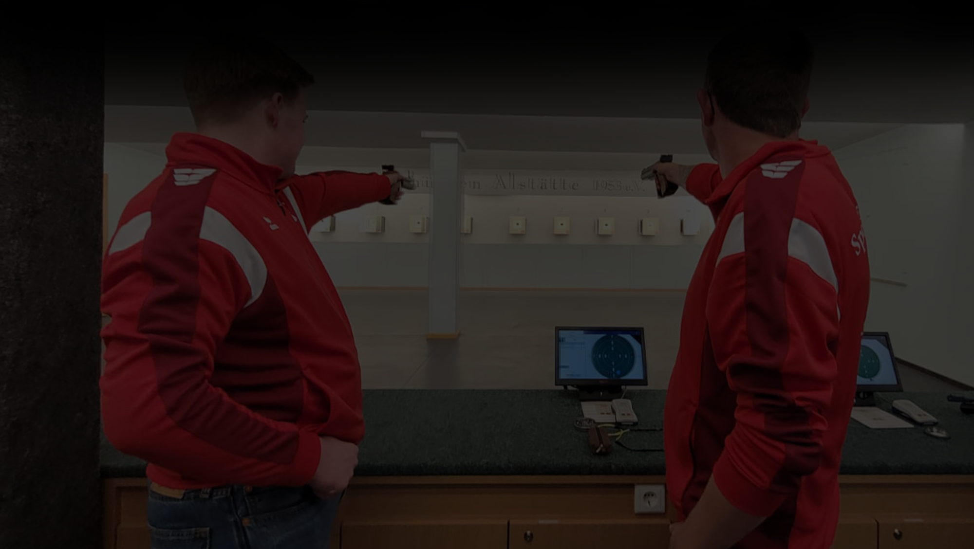 Zwei Männer zielen mit einer Schusswaffe auf eine Zielscheibe. Auf dem Bild liegt eine schwarze transparente Fläche