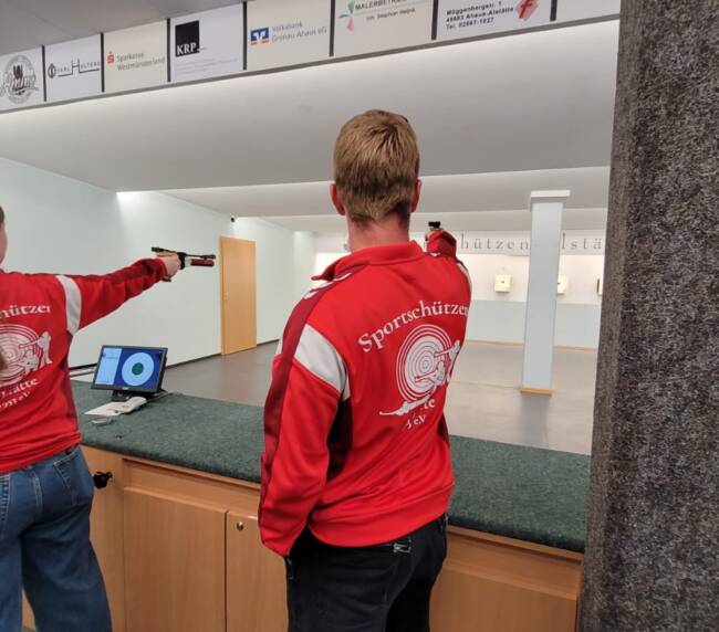 Eine Schützin und ein Schütze zielen mit einer Kurzwaffe auf eine Wand mit Zielscheiben