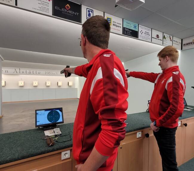 Zwei Männer sieht man seitlich von hinten. Beide Zielen mit einer Kurzwaffe auf Zielscheiben an einer Zielwand