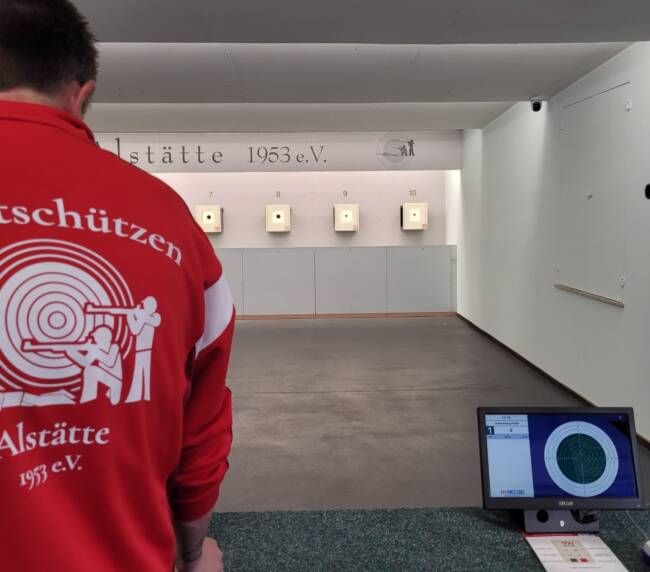 Mann steht mit dem Rücken zur Kamera und steht vor einer Zielscheiben Wand. Auf den Rücken ist das Sportschützen Logo zu sehen