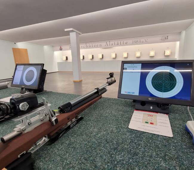 Gewähre liegen mit Bildschirmen auf denen eine Zielscheibe abgebildet sind auf einem Tisch. Im Hintergrund sind Zielscheiben an der Wand aneinandergereiht